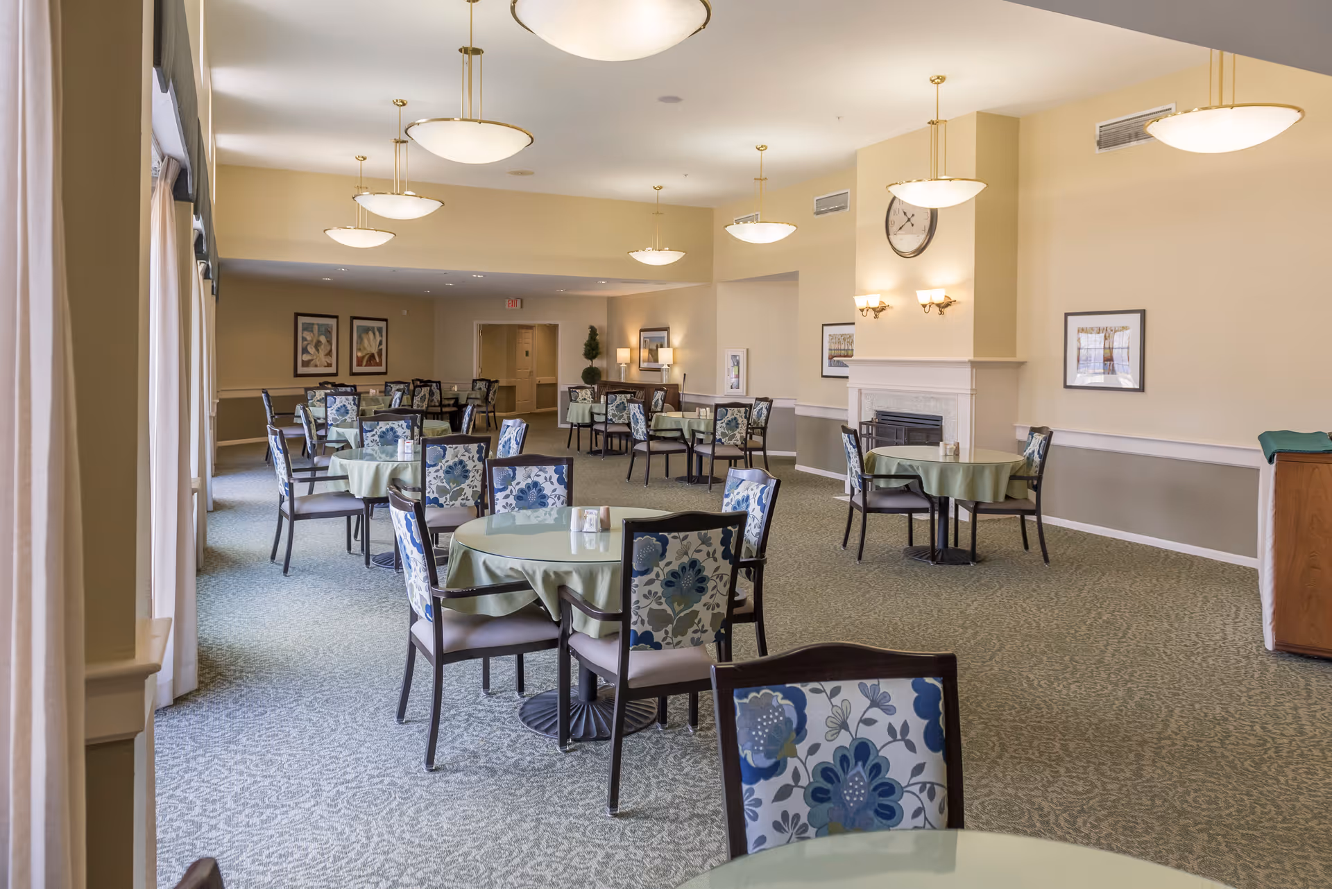 Bright dining room with round tables covered in light green tablecloths and floral-upholstered chairs arranged under pendant lights and a wall clock above a fireplace.