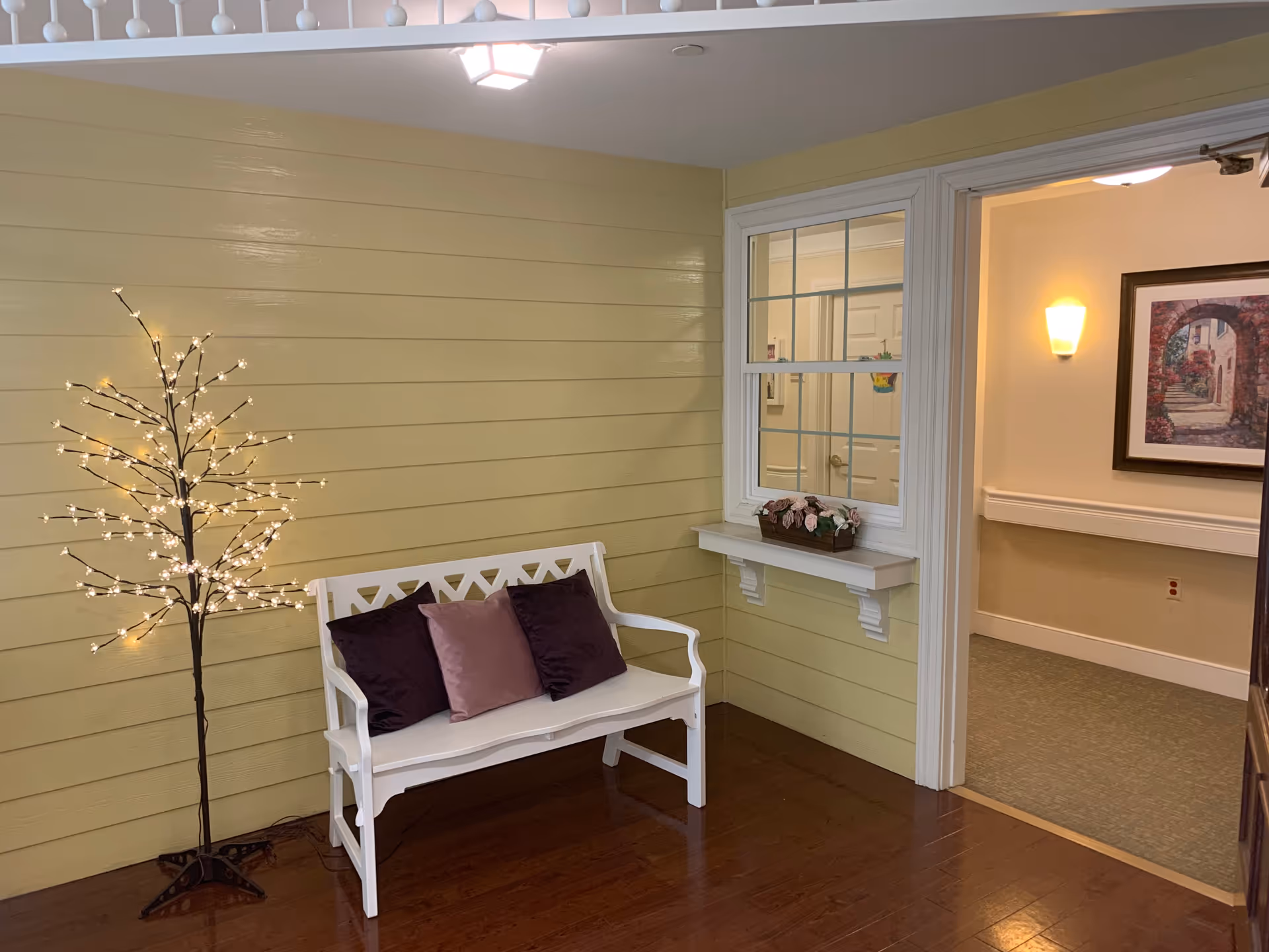 A cozy indoor seating area with a white wooden bench adorned with three pillows in dark purple and mauve. To the left of the bench is a decorative lighted tree with small white lights. The walls are painted light yellow with horizontal paneling. To the right, there is a window with a flower box containing artificial flowers and an open doorway leading to a hallway with beige walls, a wall sconce, and a framed picture.