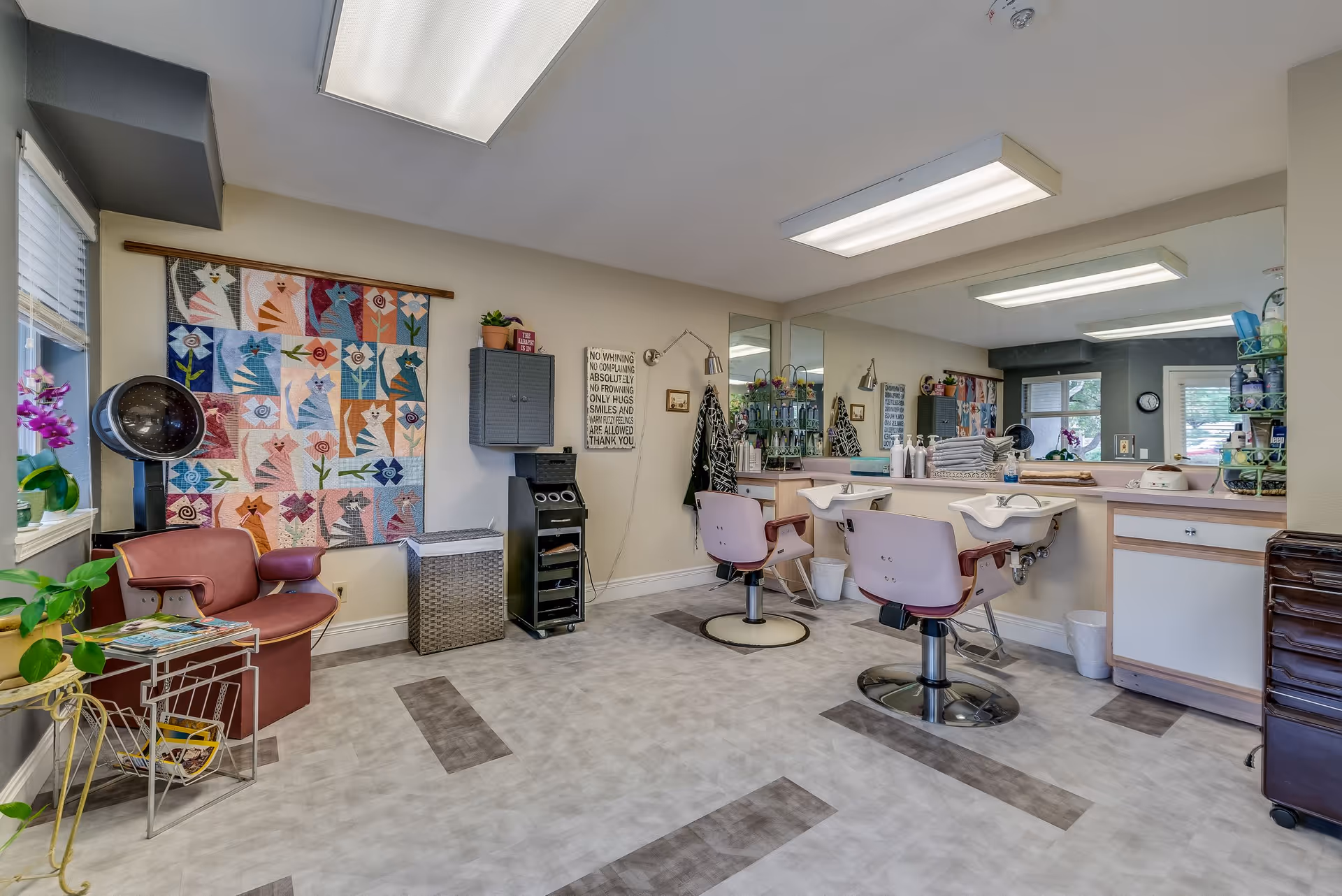 Interior view of a hair salon area in a senior living facility with two salon chairs in front of sinks and mirrors, a hair dryer chair, a colorful quilt with cat designs hanging on the wall, and various hair care products and towels on the counter.