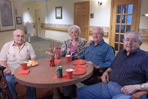 Four elderly individuals sitting around a round table with red plates, red napkins, and soda cans in a well-lit room with beige walls and wooden doors in the background.