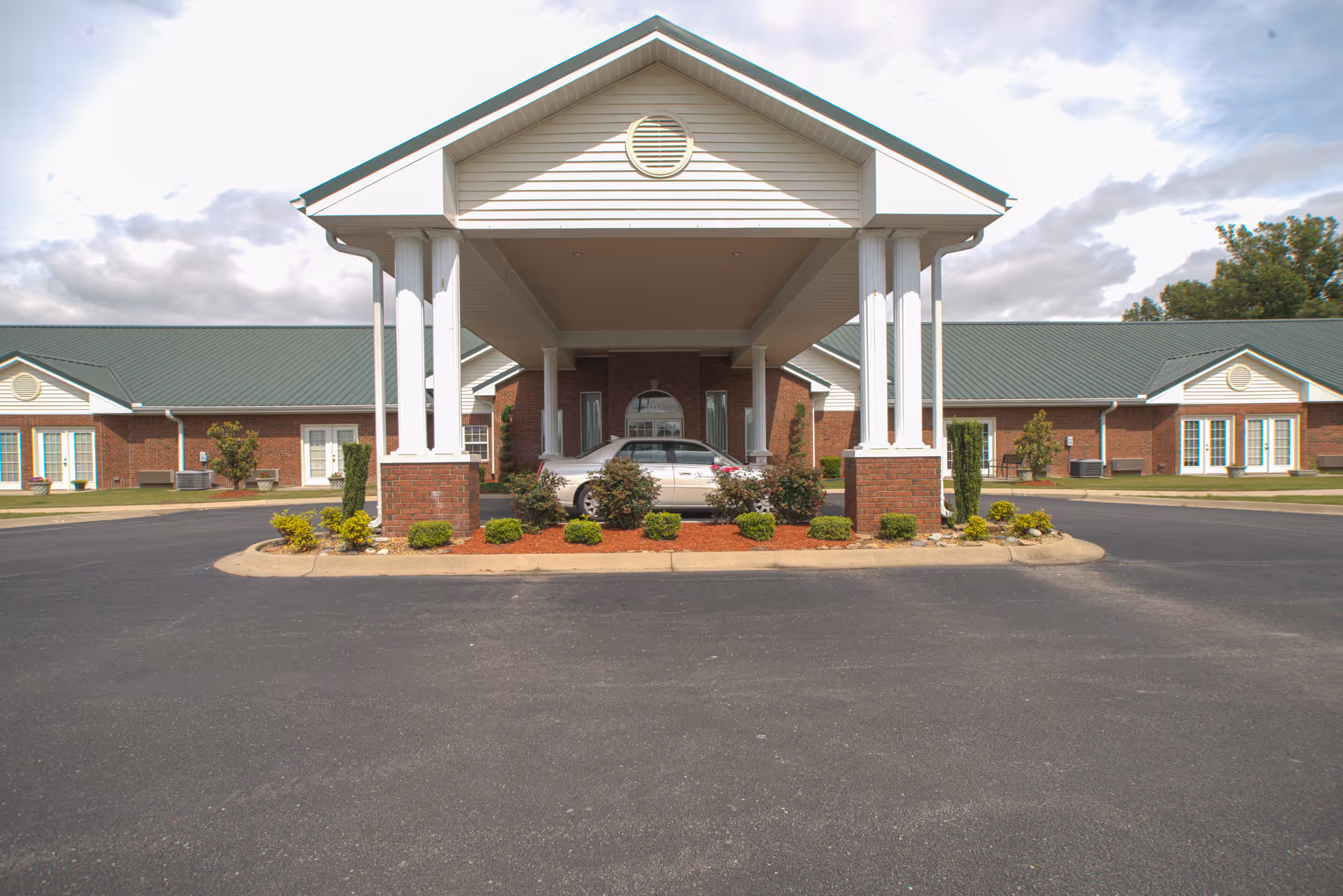 Front entrance of a single-story brick senior living building with a covered porte-cochère supported by white columns, landscaping and a parked car.