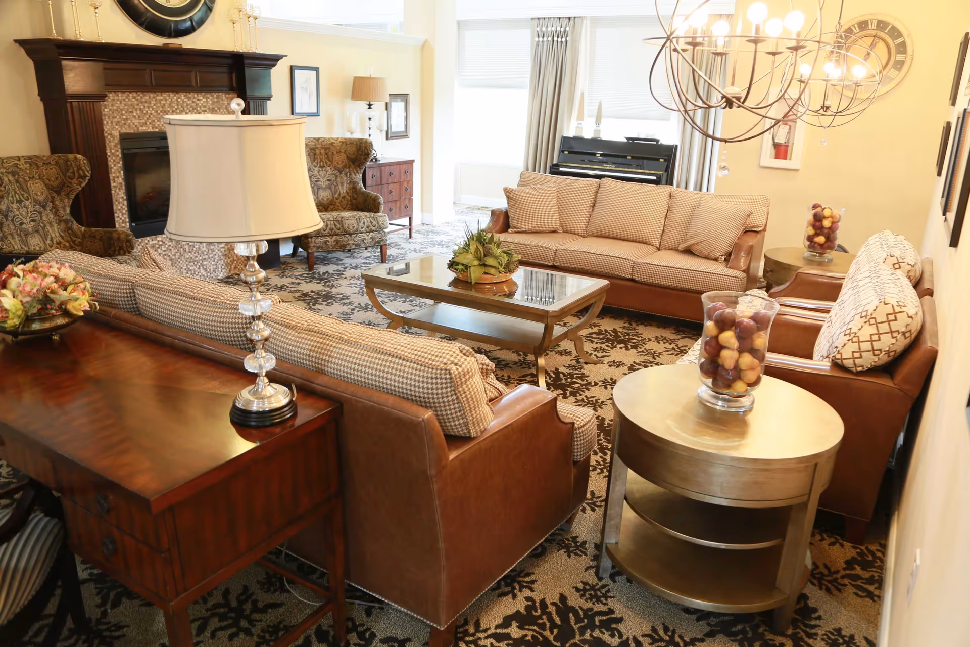 A cozy living room with two brown leather sofas, two patterned armchairs, a glass coffee table, and a wooden side table with a lamp. The room features a fireplace with a large clock above it, a patterned carpet, and a piano near the window with curtains.