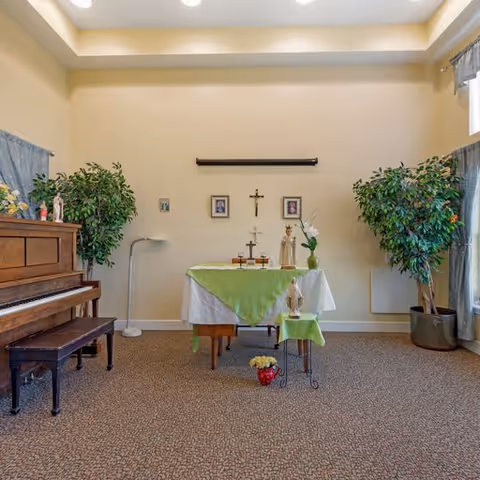A small chapel or prayer room with a piano on the left, two large potted plants on either side, and a table in the center covered with a white and green cloth holding religious items including crosses and candles. The walls are light-colored, and there are framed pictures and a crucifix hanging above the table.