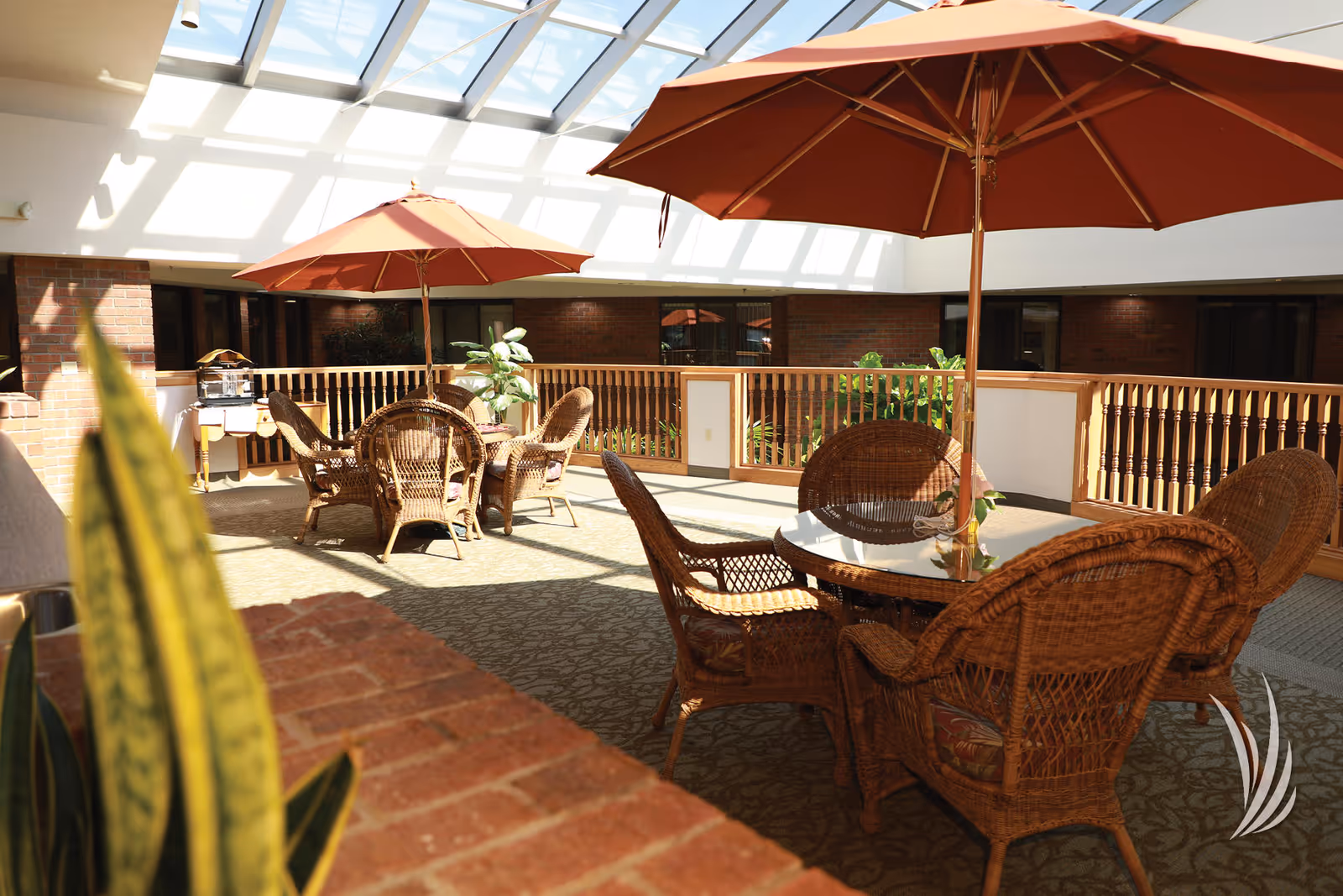 Indoor seating area with wicker chairs and round glass tables shaded by large orange umbrellas under a skylight ceiling, surrounded by brick walls and wooden railings with some plants.