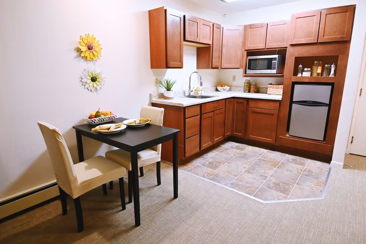A small kitchen area with wooden cabinets, a countertop with a sink, a microwave, and a mini refrigerator. Adjacent to the kitchen is a small dining table with two beige upholstered chairs, set with plates and napkins. The wall has two decorative flower hangings, and there is a basket of fruit on the table.