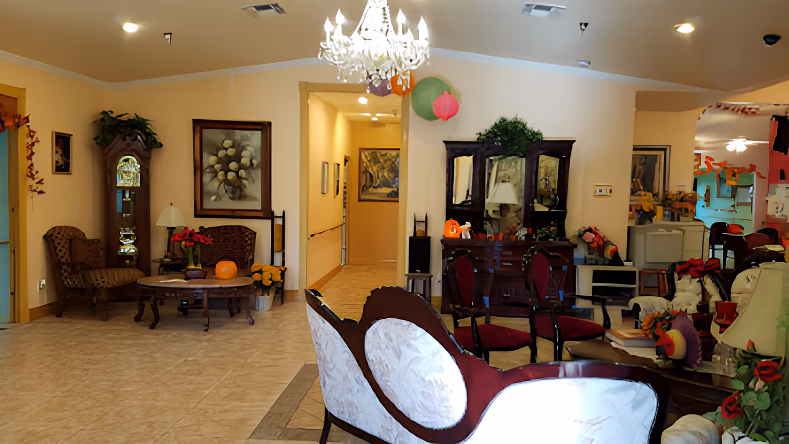 A cozy living room area in East Lake Manor Assisted Living Facility featuring vintage-style furniture including a white and maroon upholstered sofa, wooden chairs, and a coffee table with a pumpkin decoration. The room has warm beige walls, a chandelier hanging from the ceiling, framed artwork, plants, and a wooden cabinet with a mirror. There is a hallway visible in the background.