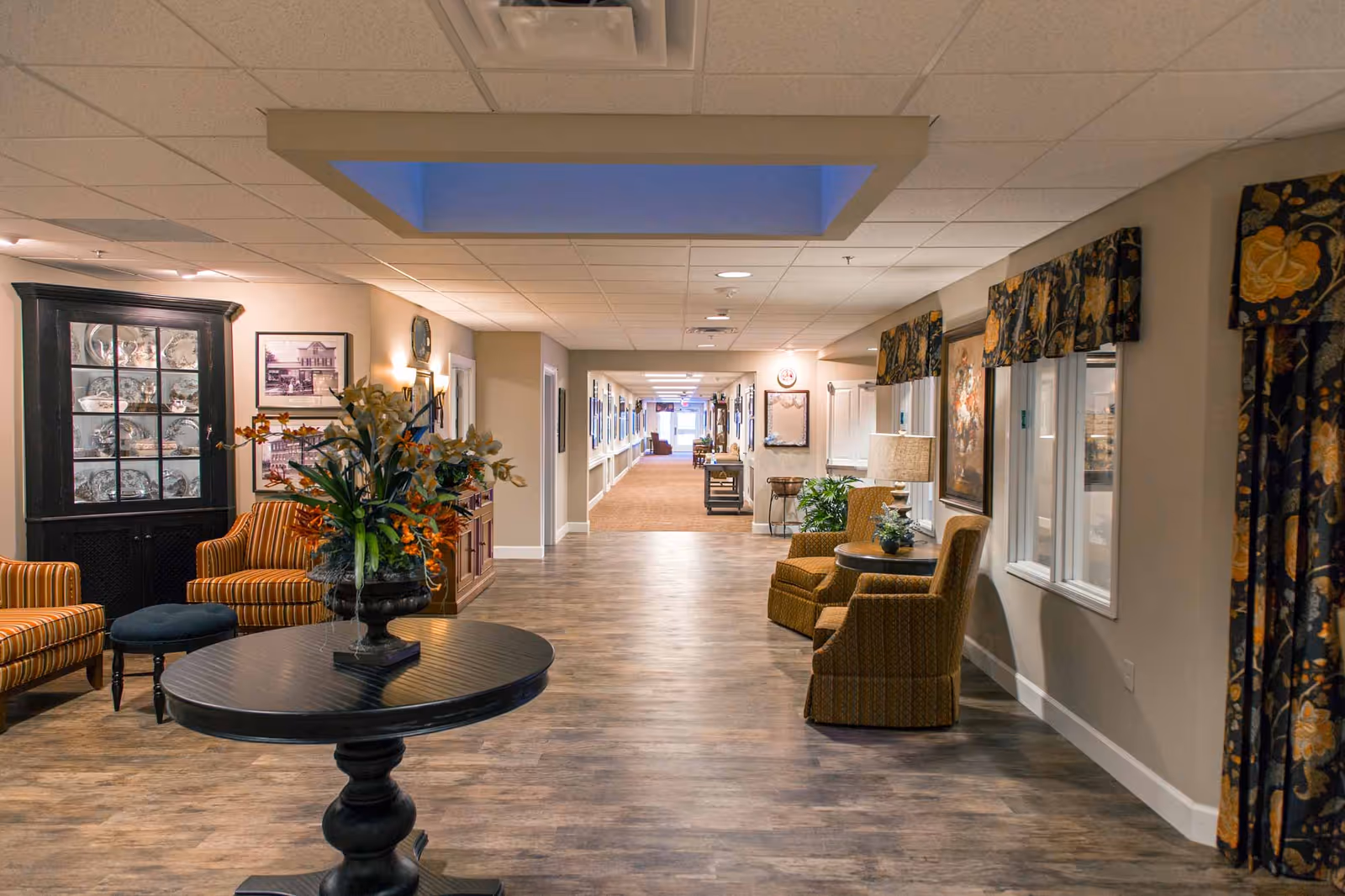 A well-lit hallway in a senior living facility with wood flooring and beige walls. The hallway features comfortable upholstered chairs with patterned fabric, a round black table with a floral arrangement, framed pictures on the walls, and windows with floral valances. The corridor extends into the distance with additional seating and decor visible.