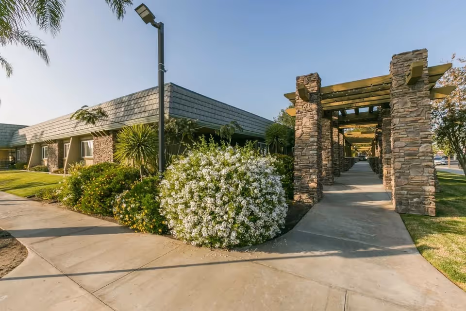 Stone-column pergola walkway next to a single-story building with landscaped shrubs and a sidewalk.