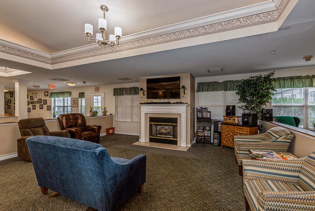 A cozy living room area in a senior living facility featuring a blue upholstered loveseat, two patterned armchairs, and two brown recliners arranged around a fireplace with a mounted flat-screen TV above it. The room has carpeted floors, green valance curtains on the windows, a chandelier hanging from the ceiling, and various decorative items including plants and framed pictures on the walls.