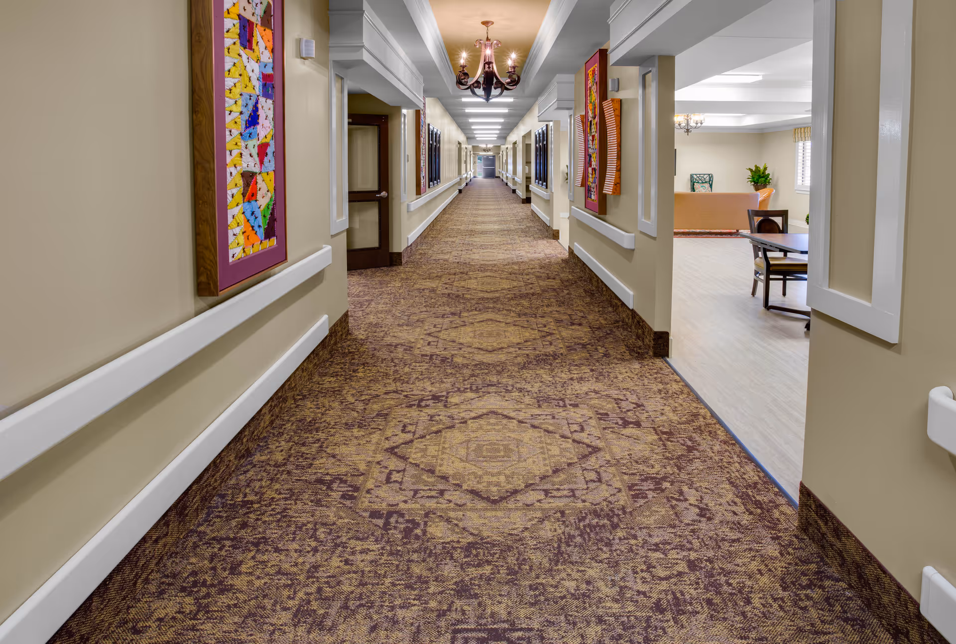 A long carpeted interior hallway in a senior living facility with handrails, framed artwork, chandeliers, and an open seating area to the right.