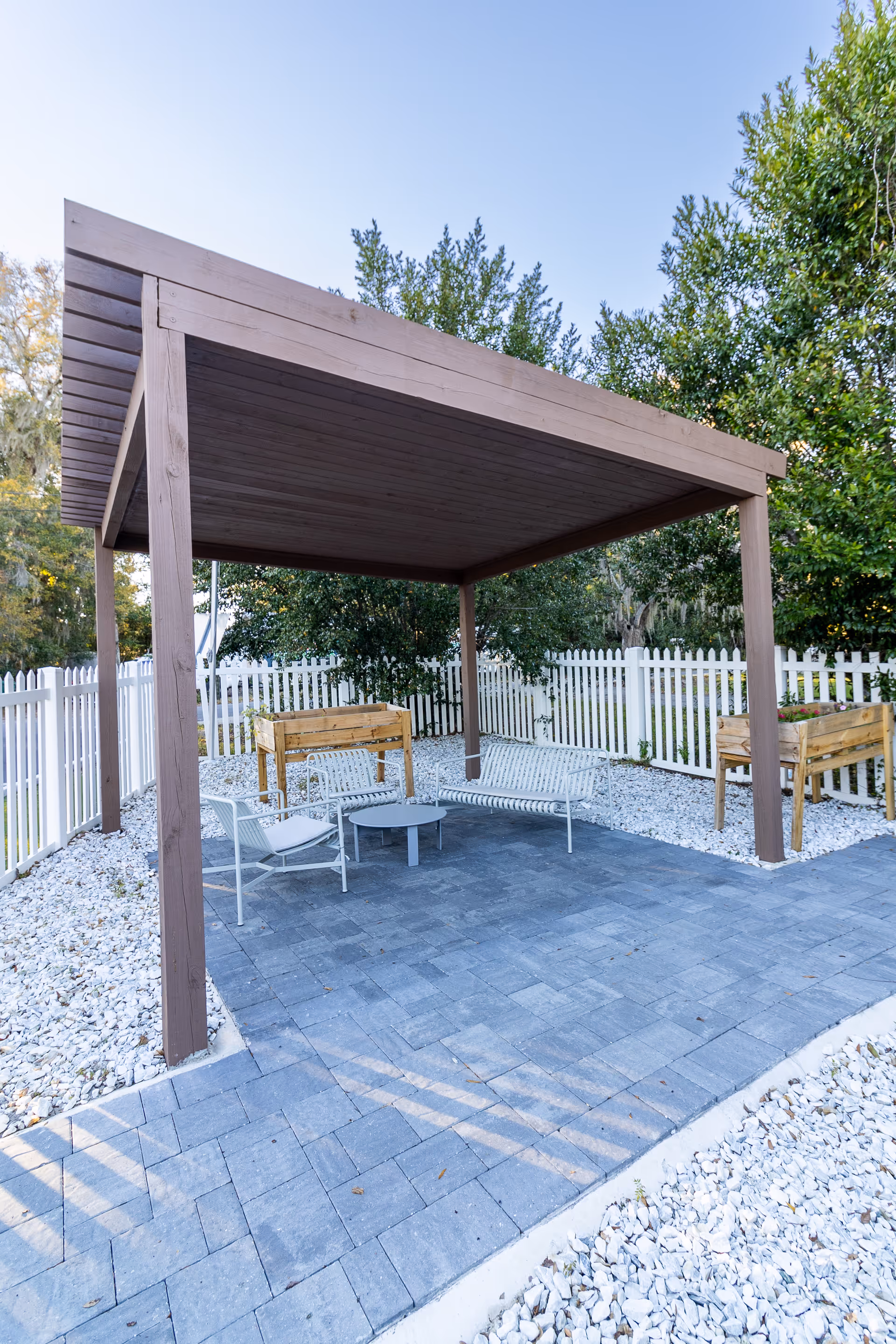 Outdoor patio area with a wooden pergola providing shade over a seating arrangement consisting of two white metal chairs, a white metal bench, and a small round table. The patio is paved with gray stone tiles and surrounded by white gravel and a white picket fence. There are two wooden planter boxes with plants on either side of the pergola, and green trees and bushes are visible in the background.