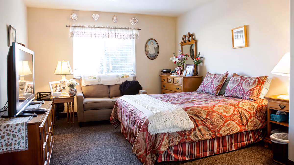 Cozy furnished bedroom with a double bed in patterned bedding, a sofa by a window, dresser, TV, and bedside tables with lamps.