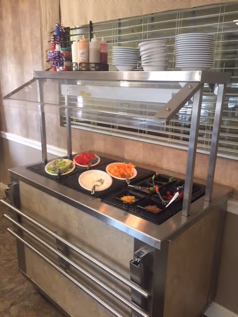 Self-serve buffet station with bowls of salad, condiments and stacks of plates under a sneeze guard.