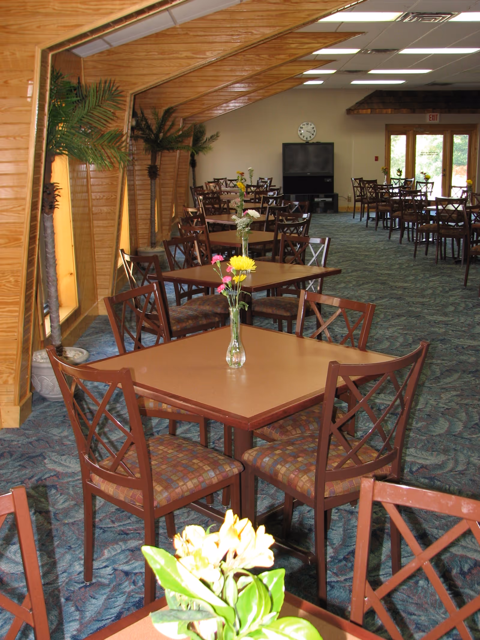 Dining room with square tables and chairs, floral centerpieces, wood-paneled walls and a TV at the far end.