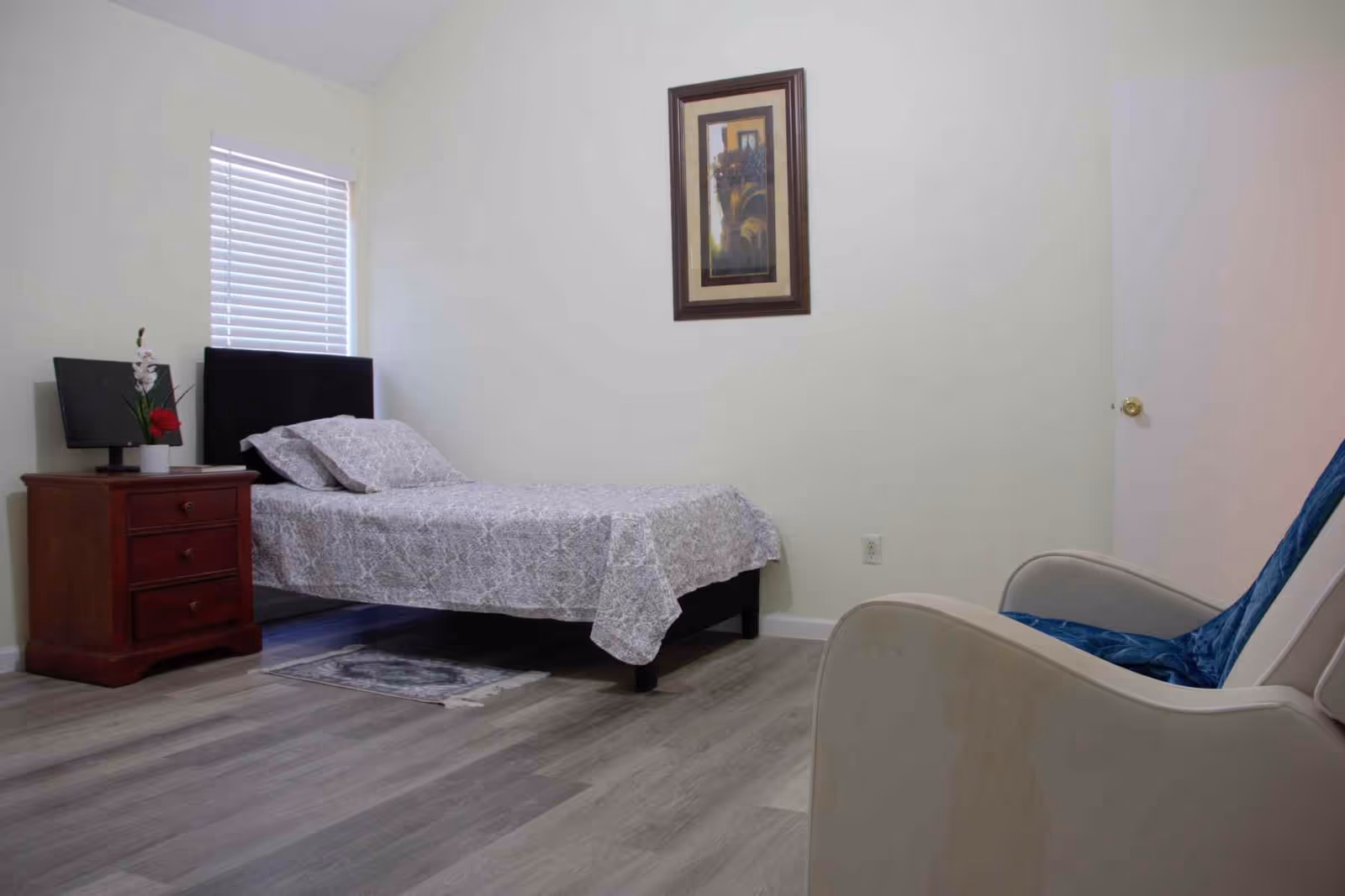 A simple bedroom with a single bed covered in a patterned gray bedspread, a wooden nightstand with a small plant and a monitor on it, a framed picture hanging on the wall, a window with closed blinds, and a beige armchair with a blue throw draped over it.