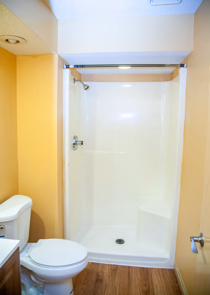 A small bathroom with a white toilet and a white shower stall with a built-in seat. The walls are painted yellow, and the floor has wood-like vinyl flooring. A silver showerhead and control knob are visible inside the shower.