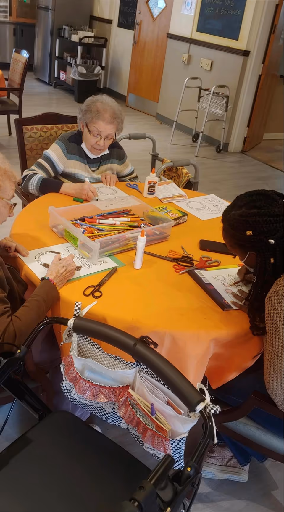 Three people sitting around a table covered with an orange tablecloth, engaged in coloring activities with crayons, markers, glue, and scissors. Two elderly women and one younger woman are coloring on paper. A walker with a fabric organizer is in the foreground. The room has a kitchen area and a walker in the background.