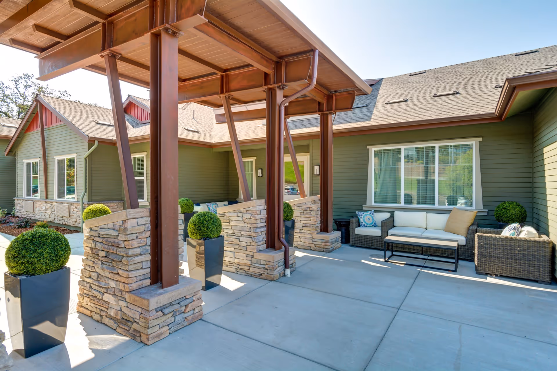 Outdoor patio area at The Vineyard at Fountaingrove Memory Care featuring a covered entrance with stone pillars and brown wooden beams, surrounded by green plants in tall black planters. There is a seating area with a wicker sofa and chairs with cushions and pillows, set against the green exterior walls of the building under a clear blue sky.