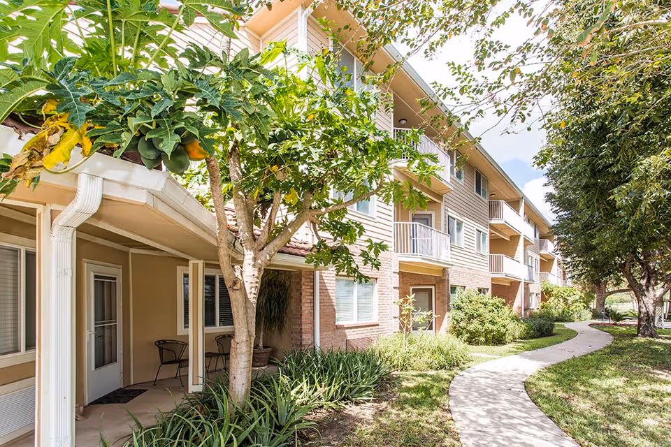 Exterior view of a multi-story retirement community building with balconies, surrounded by trees and greenery. A curved concrete pathway leads through the landscaped garden area in front of the building.
