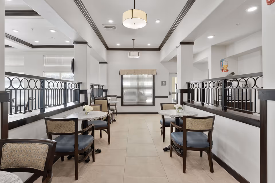A bright and clean dining area with small round tables and cushioned chairs arranged along a corridor. The space features white walls, large windows with blinds, and decorative black railings separating the dining area from an adjacent space. Ceiling lights hang above the tables, and small flower arrangements are placed on each table.
