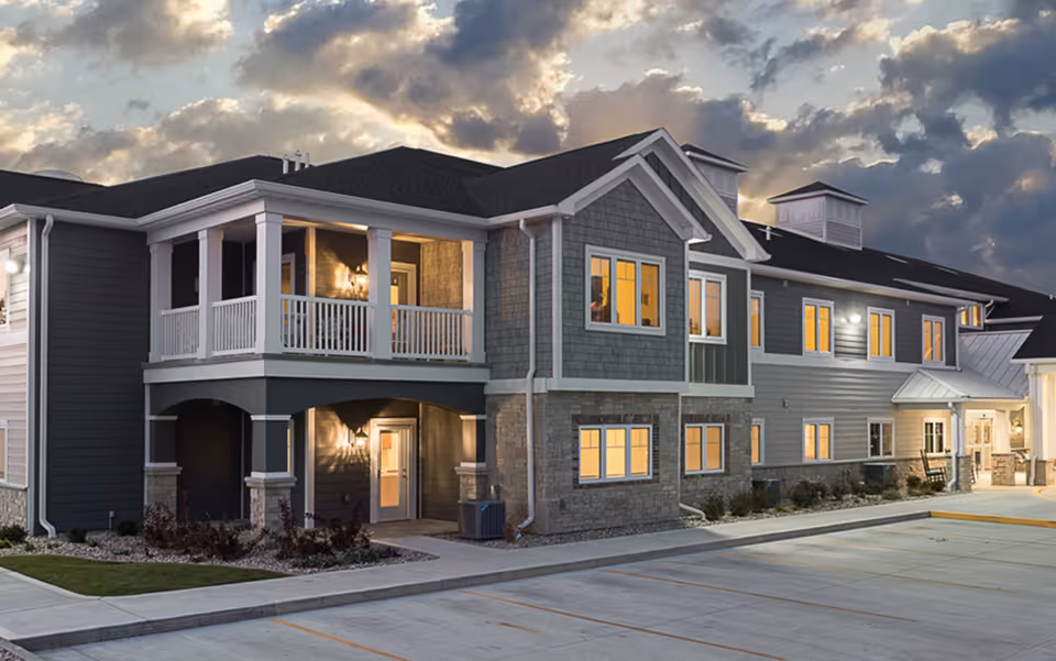 Exterior view of a modern two-story senior living facility building at dusk with lit windows, a balcony, and a parking area in front under a cloudy sky.