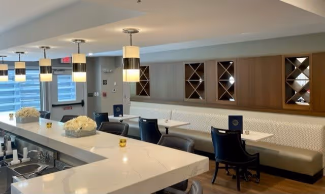 A modern dining area in a senior living facility featuring a long white marble countertop with bar stools, pendant lights hanging from the ceiling, a row of tables with black chairs along a wall with built-in wooden wine racks, and decorative flower arrangements on the counter.