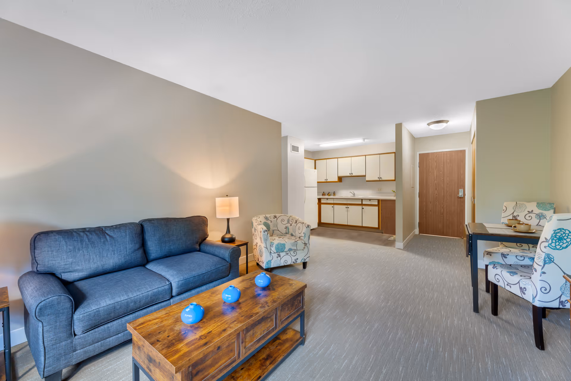 Bright furnished living area with a blue sofa, wooden coffee table, patterned chairs, and a kitchenette in the background.