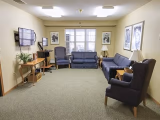 A cozy living room with beige walls and carpeted floor, featuring two striped blue sofas, two matching armchairs, a wooden side table with a lamp, a wooden console table with a plant and electronic equipment, and framed black and white portraits on the walls. A large window lets in natural light.