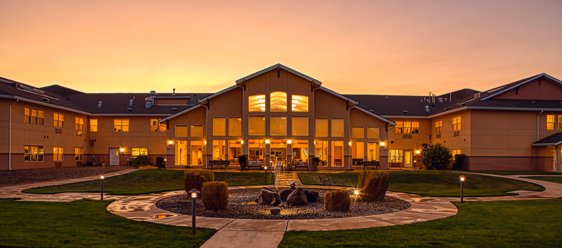 Exterior view of Wheatfields Estates Senior Living and Memory Care building at sunset, showing a two-story structure with large windows illuminated from inside, surrounded by a landscaped garden with pathways and small bushes.