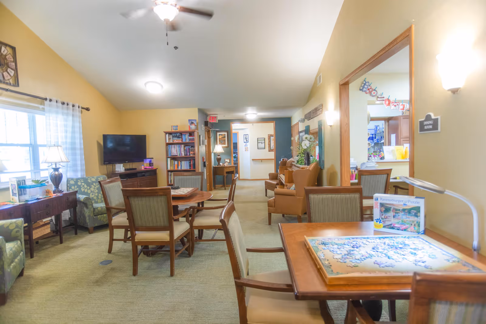 Bright communal sitting and activity room with tables, chairs, armchairs, a TV, bookshelves and a puzzle on a table.