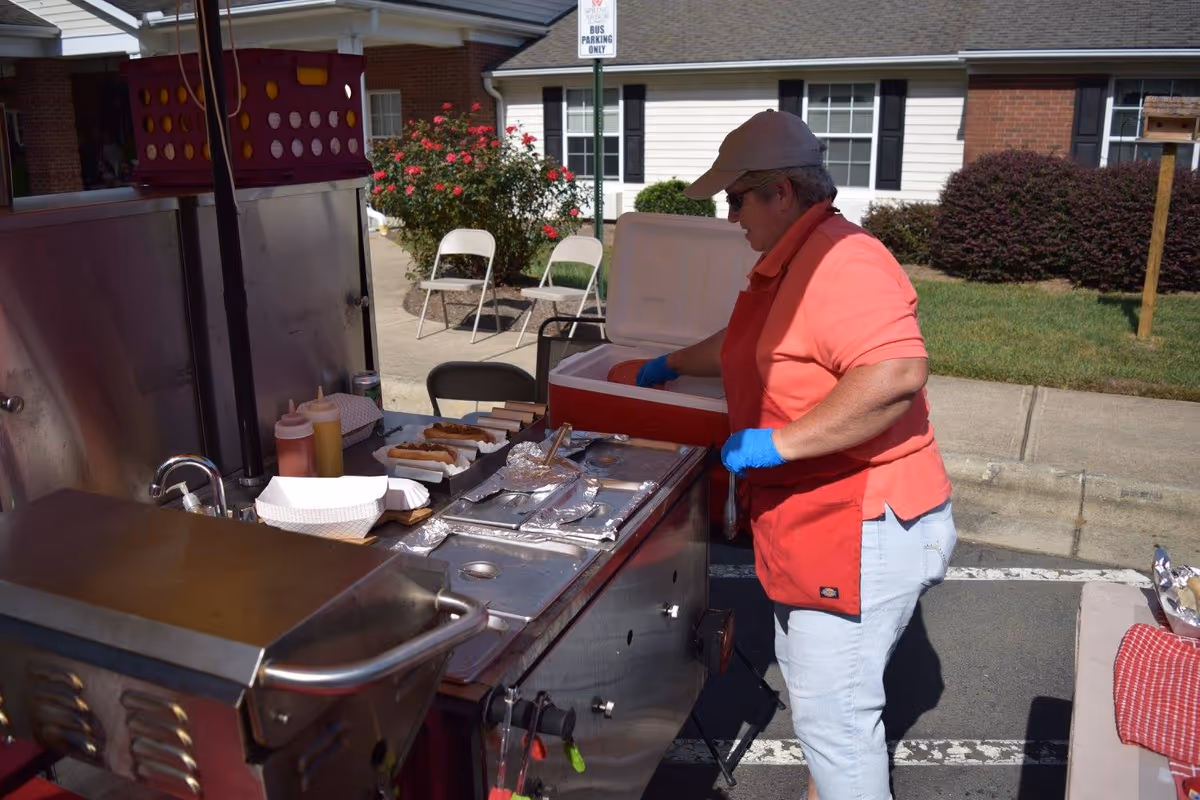 A person wearing a red shirt, white pants, a beige cap, and blue gloves is preparing food at an outdoor grill station. There are hot dogs and condiments on the grill counter. In the background, there are white folding chairs, a bush with red flowers, and a building with white siding and brick accents.