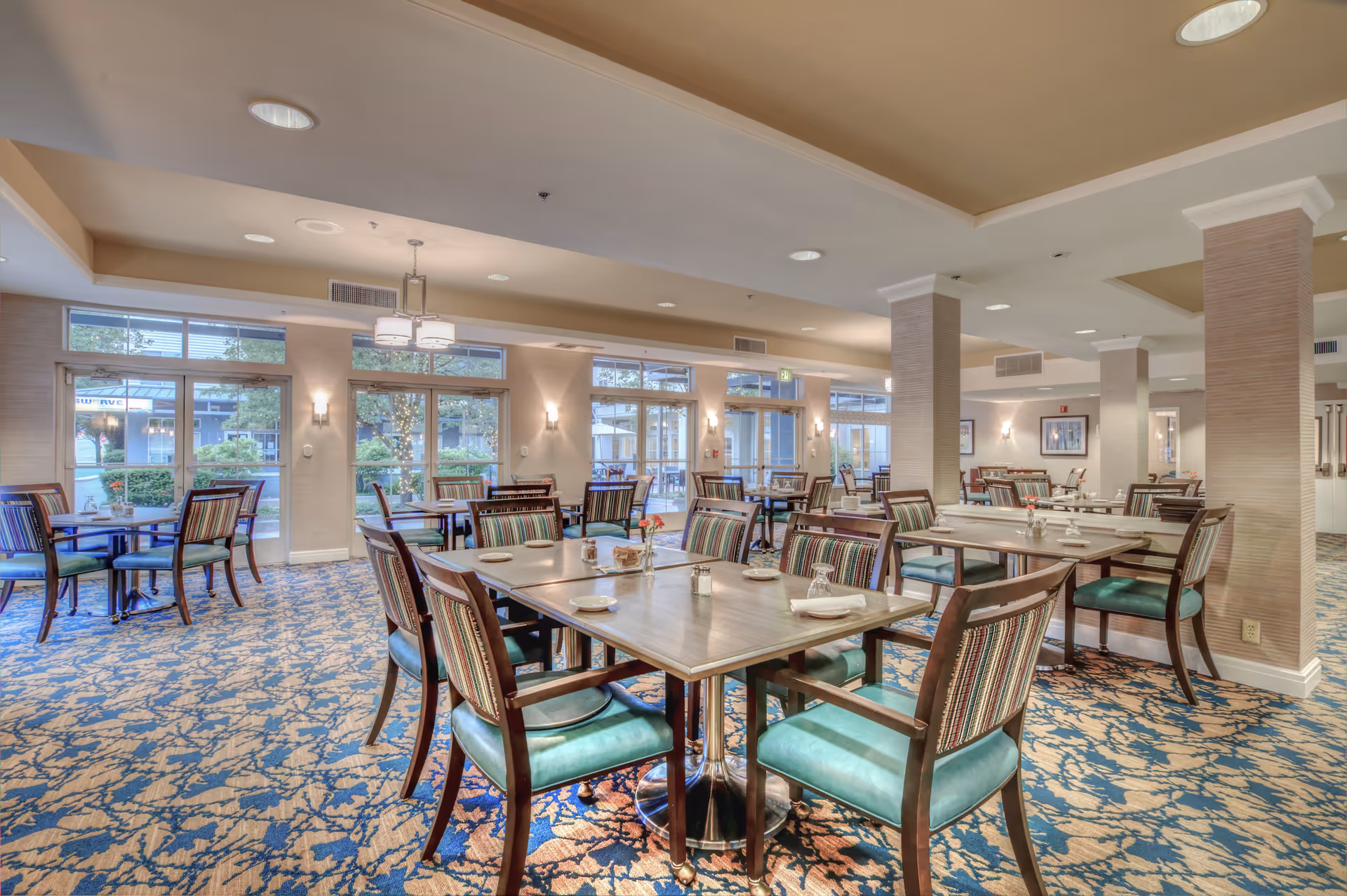 Bright, spacious dining room with multiple wooden tables and upholstered chairs and large windows overlooking greenery.