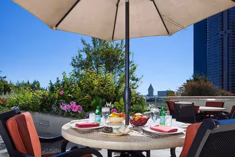 Outdoor rooftop dining table with umbrella, chairs, flowers, and distant city skyline.
