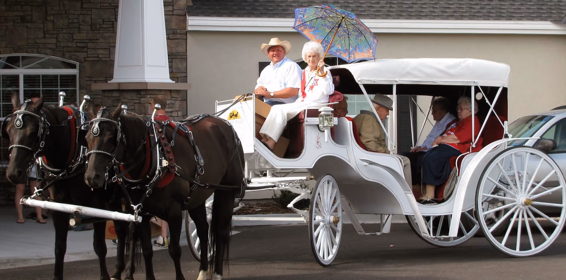 A group of elderly people riding in a white horse-drawn carriage with two black horses in front. One elderly woman is holding a colorful umbrella, and a man wearing a cowboy hat is sitting beside her. The carriage is parked outside a building with stone and beige walls.