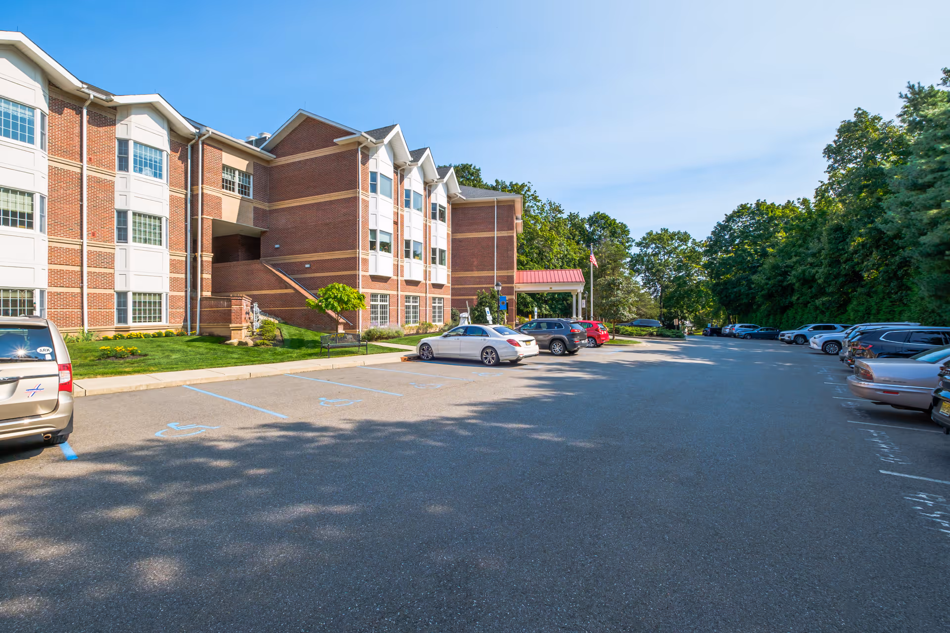 Parking lot in front of a multi-story brick building with white window frames and a covered entrance. Several cars are parked along the sides of the lot, and there are trees and greenery surrounding the area under a clear blue sky.