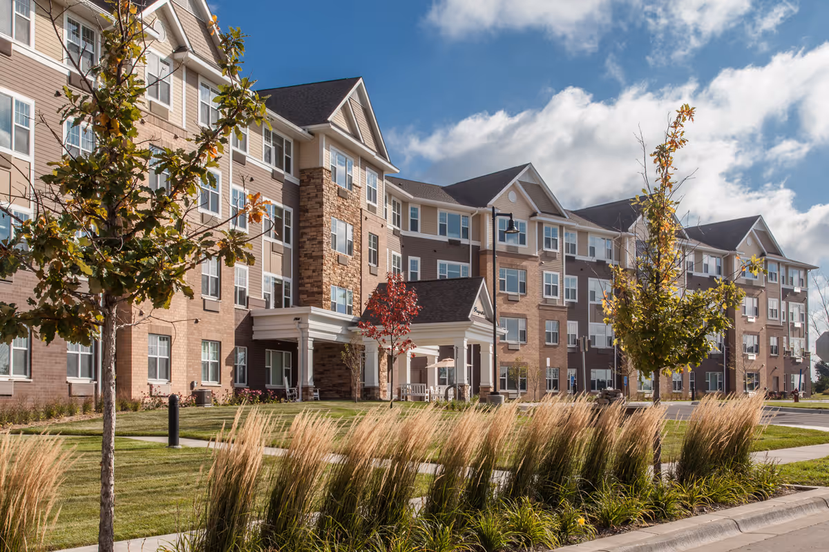 Exterior view of a multi-story senior living facility building with a mix of brick and siding, surrounded by landscaped greenery including trees and ornamental grasses under a partly cloudy sky.