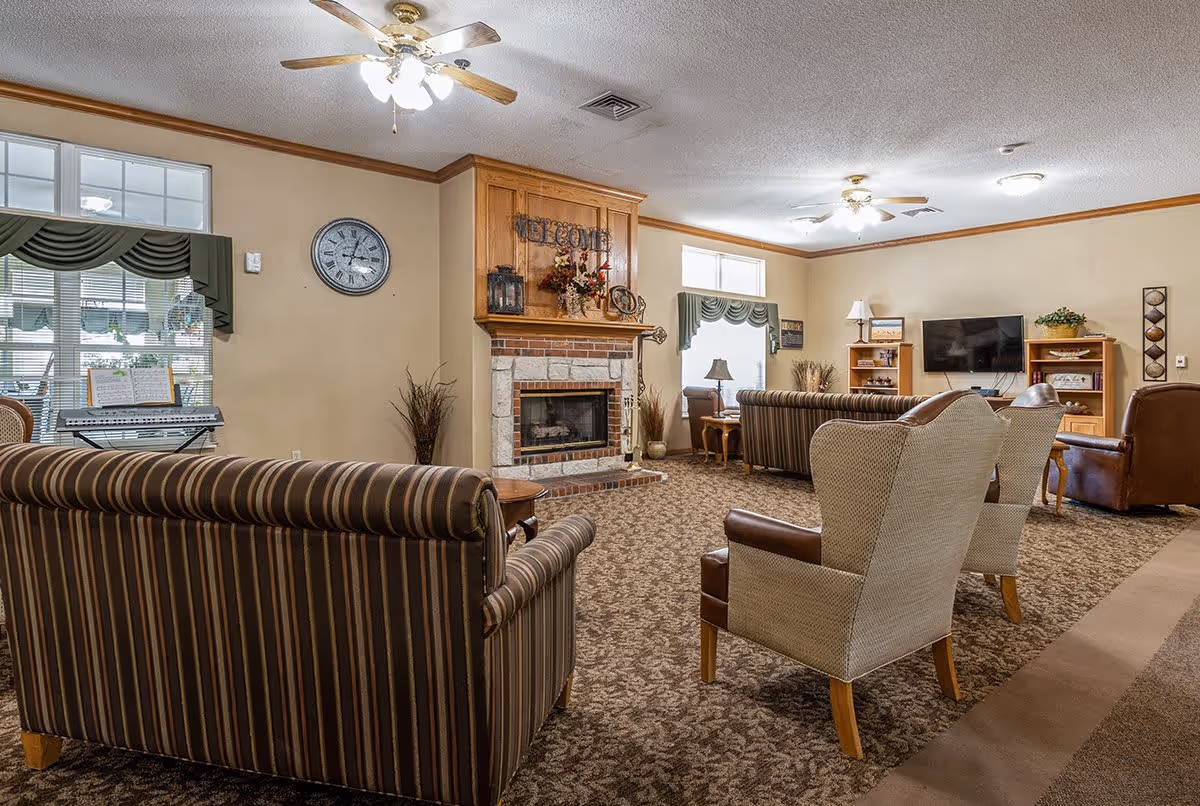 A cozy living room with patterned carpet, striped sofas, and beige armchairs arranged around a brick fireplace with a wooden mantel decorated with flowers and a welcome sign. The room has ceiling fans with lights, windows with green valances, a wall clock, a keyboard near the window, and a TV mounted on the wall with wooden shelves on either side.