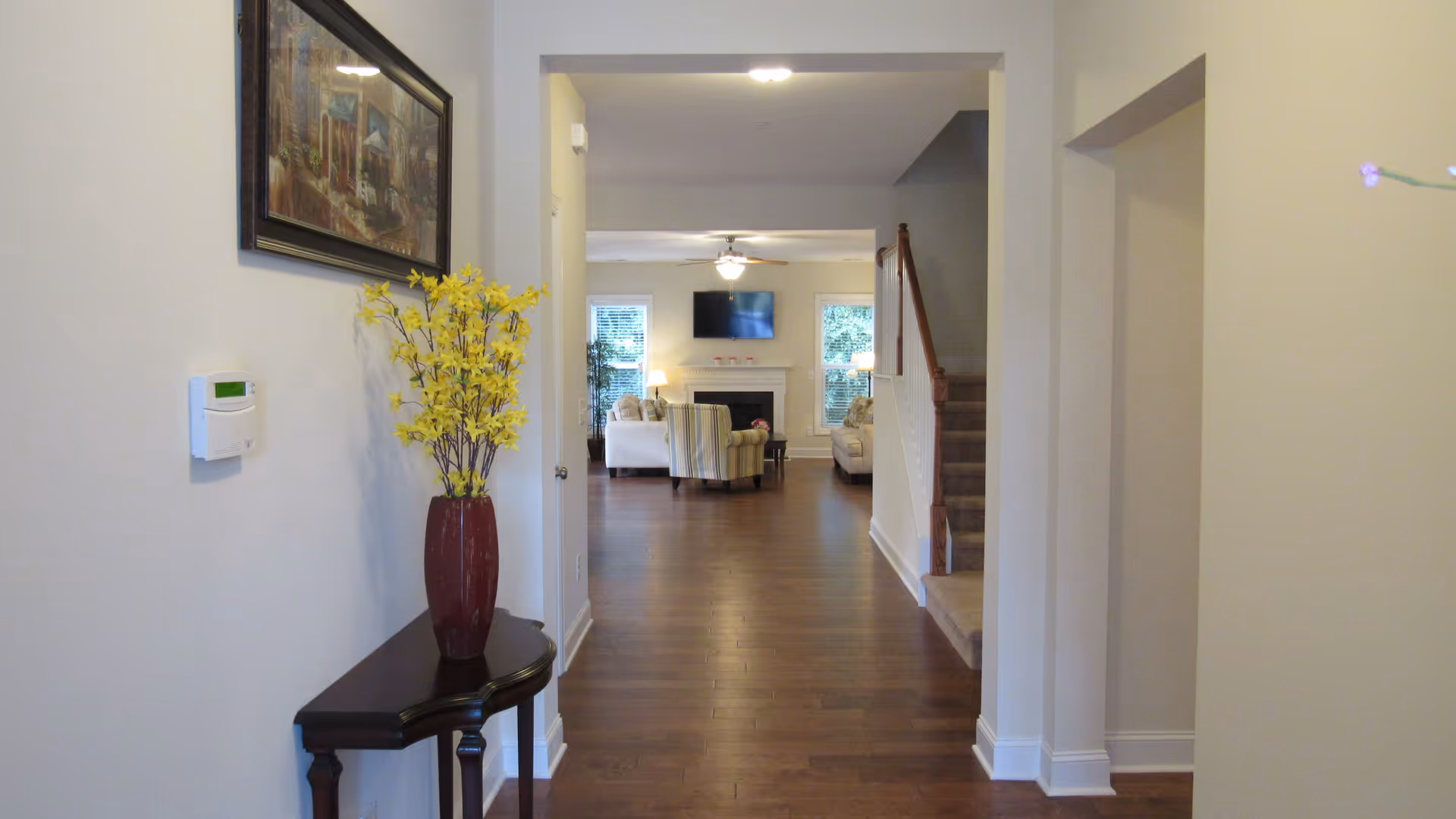 View down a hallway with wooden flooring leading to a living room area. On the left side of the hallway, there is a small dark wooden table with a tall red vase holding yellow flowers. A framed painting hangs above the table. The living room has two sofas, an armchair, a fireplace with a TV mounted above it, and two windows with blinds. A staircase with carpeted steps and a wooden handrail is on the right side of the hallway.