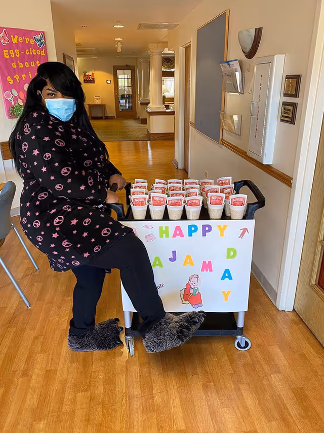 A woman wearing a face mask, black pajama top with pink peace signs and stars, black pants, and fuzzy slippers is sitting on a cart filled with cups of Nestle Nesquik. The cart has a sign that says 'Happy Pajama Day' with colorful letters and a small illustration of a child in pajamas. The setting appears to be a hallway in a senior living facility with wooden floors and light-colored walls.