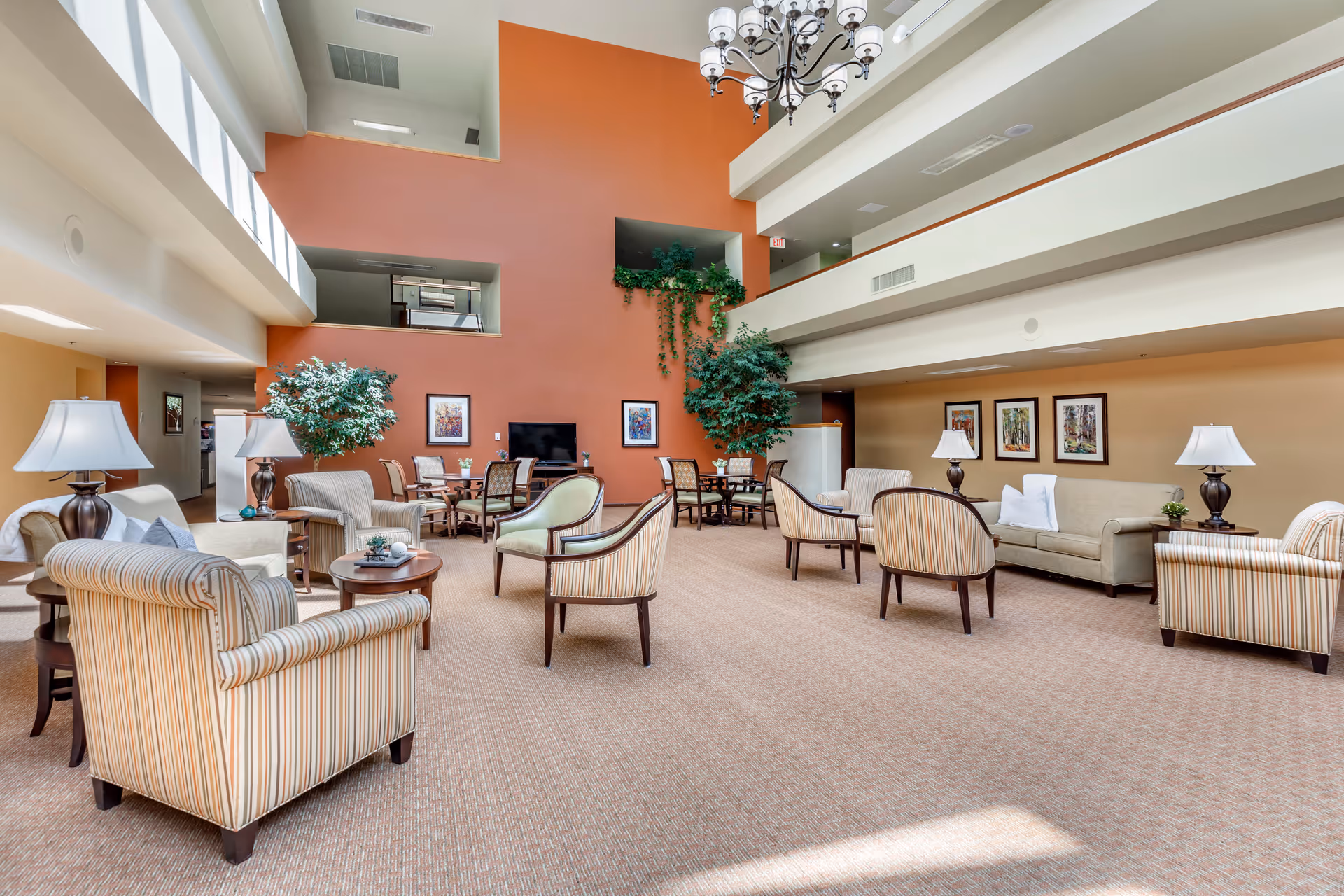 A spacious and well-lit living room area in a senior living facility with high ceilings and large windows. The room features multiple upholstered armchairs and sofas arranged around small tables, several table lamps, framed artwork on the walls, and potted plants. A chandelier hangs from the ceiling, and there is a television mounted on a burnt orange accent wall.