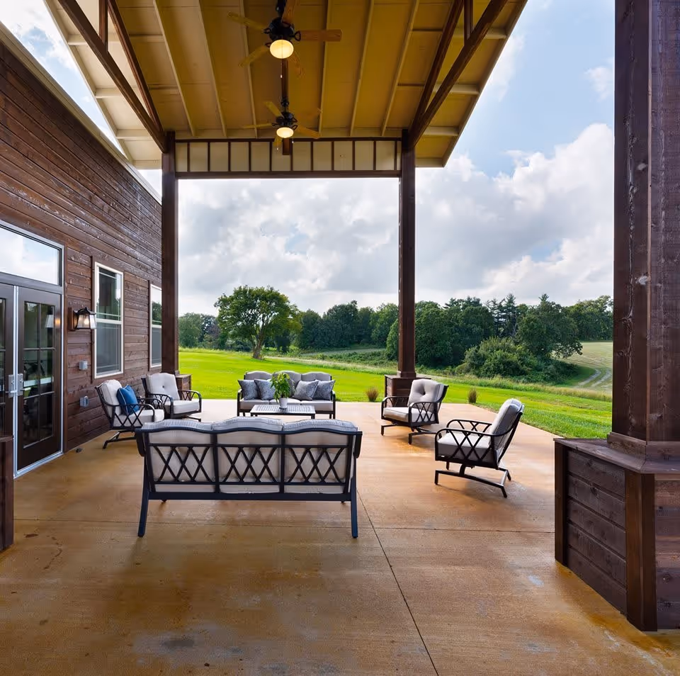 Covered outdoor patio with cushioned chairs and sofas arranged around a coffee table overlooking a grassy landscape.