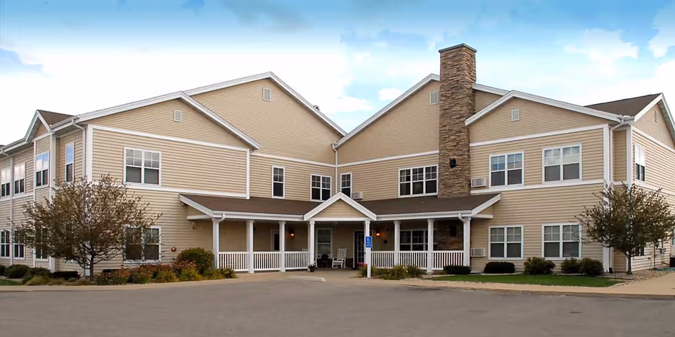 Exterior view of a two-story beige senior living facility building with white trim, multiple windows, a central covered entrance with a porch, and a stone chimney. There are small trees and shrubs around the building and a paved driveway in front.