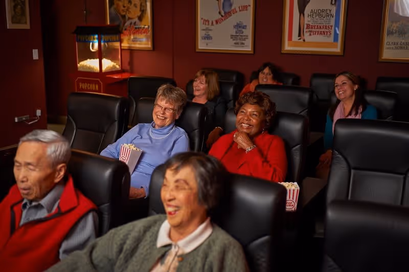 A group of elderly people sitting in comfortable black leather chairs in a small movie theater room, smiling and enjoying a film. Some are holding popcorn, and movie posters are visible on the wall behind them.