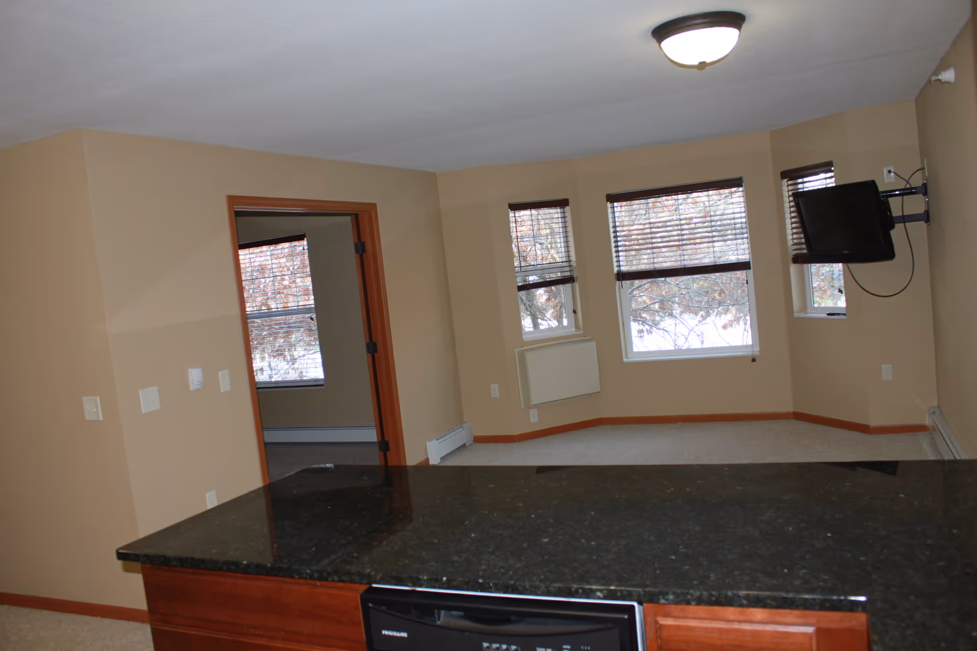Interior view of a senior living facility showing a room with beige walls, three windows with blinds, a wall-mounted TV, and a black granite countertop in the foreground. There is an open doorway leading to another room with a window.