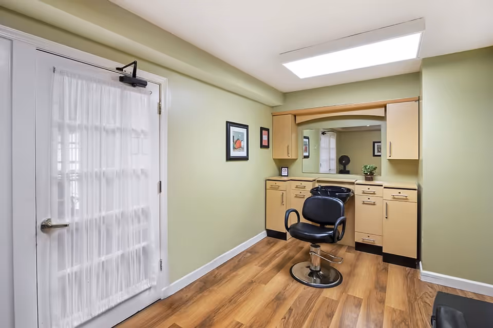 A small salon area with a black salon chair in front of a large mirror and beige cabinetry. The room has light green walls, wood flooring, and a white door with a sheer curtain.