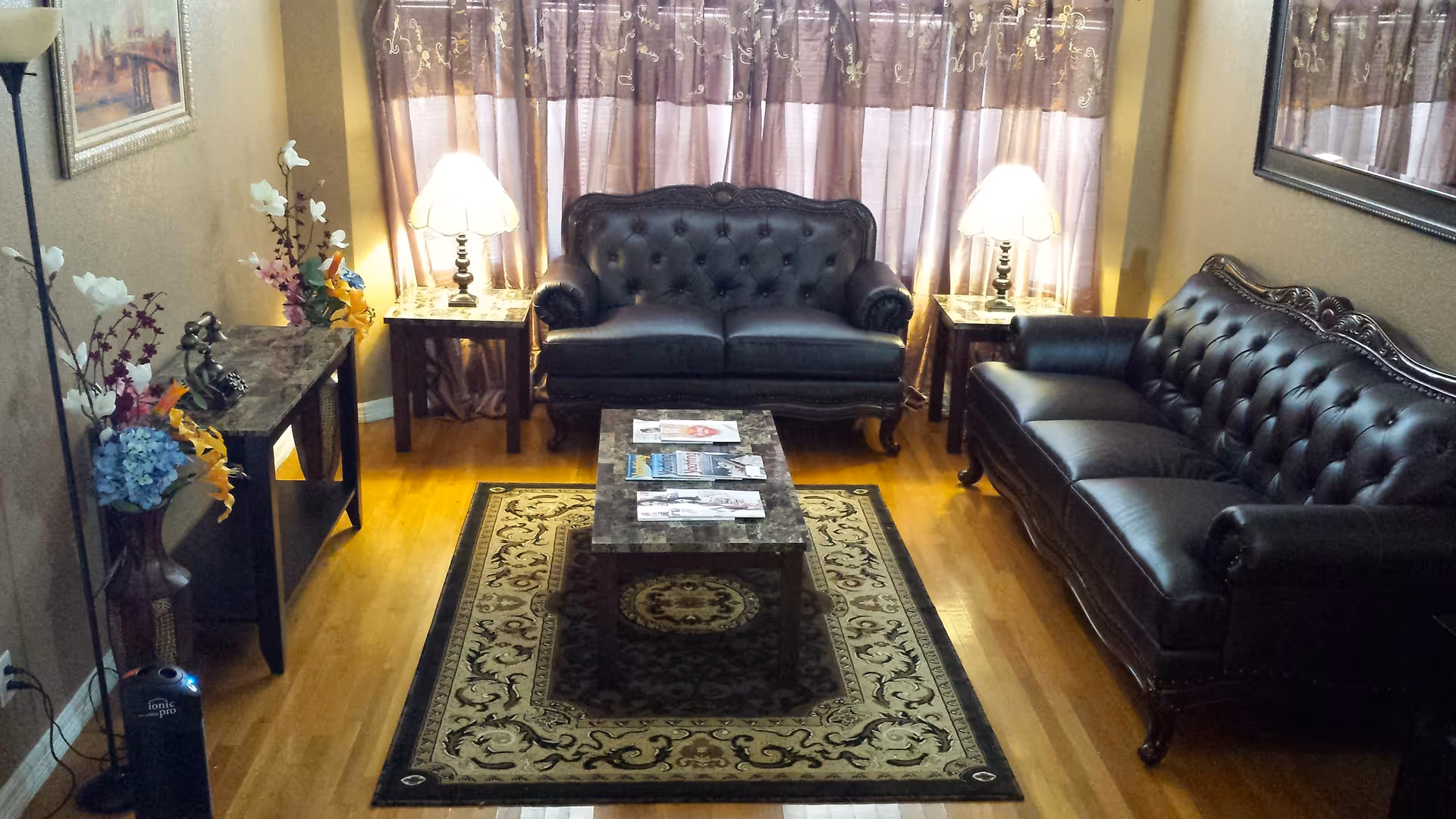 A cozy living room with two dark leather tufted sofas facing each other, a rectangular coffee table with magazines on it, two side tables each with a lamp, a patterned area rug on a wooden floor, floral arrangements on a console table, and a large window with sheer curtains in the background.