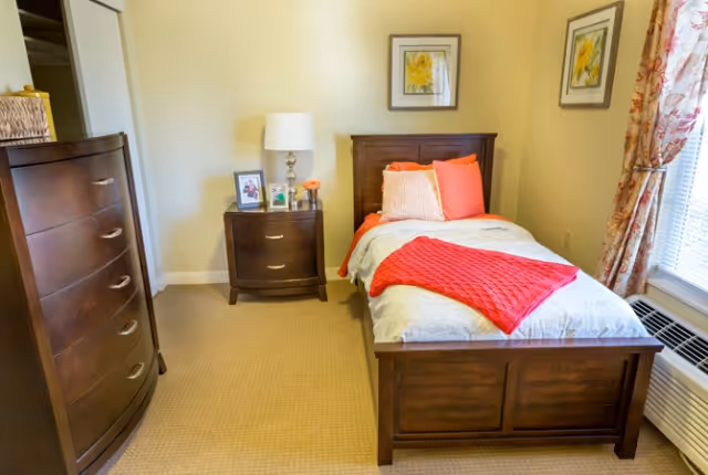A neatly arranged bedroom with a single wooden bed featuring white and coral bedding, a coral throw blanket, and matching pillows. Next to the bed is a wooden nightstand with a lamp, framed photos, and a small flower vase. A wooden dresser is positioned on the left side of the room. The room has beige walls, carpeted floor, floral curtains, and a window air conditioning unit.