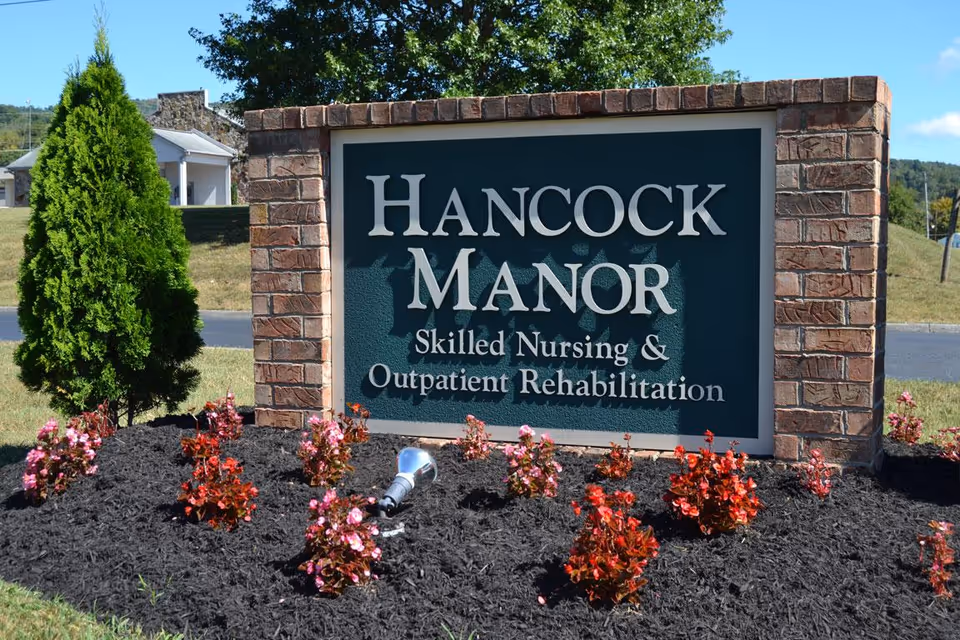 Brick entrance sign reading "Hancock Manor Skilled Nursing & Outpatient Rehabilitation" in a landscaped flowerbed with shrubs.