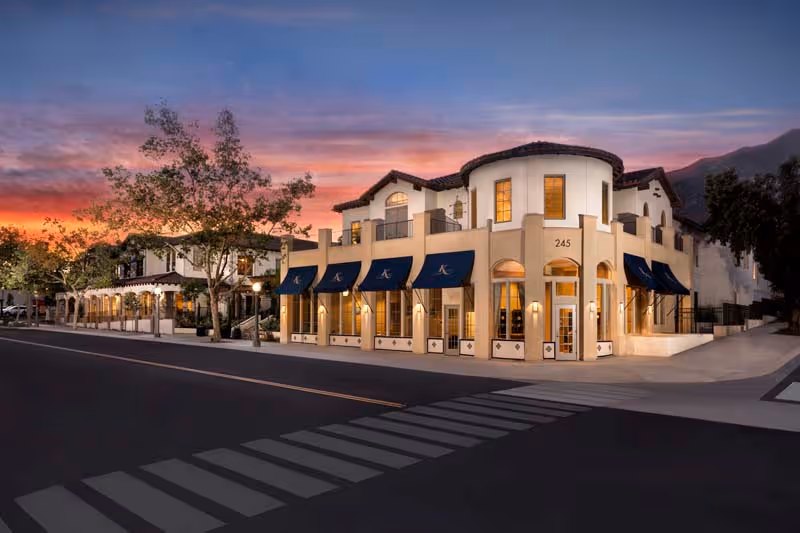 Exterior view of The Kensington Sierra Madre building at dusk, featuring a corner entrance with the number 245, multiple windows with blue awnings, and a tree-lined street with a crosswalk in the foreground.