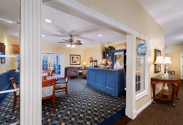 Interior view of a senior living facility common area with patterned carpet, a blue reception or snack counter with a model sailboat on top, several wooden chairs and tables, armchairs, a ceiling fan with lights, framed artwork on the walls, and a side table with a lamp and decorative items.